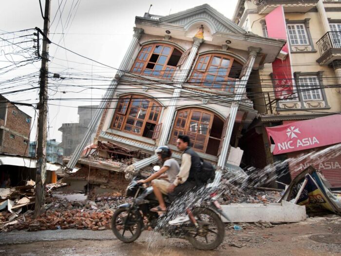 two young men on a motorbike passing a leaning devastated house